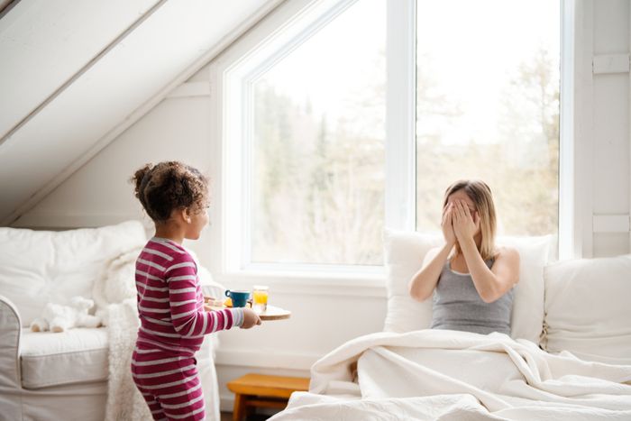 A woman sits in bed with her face covered, as her daughter carries a plate of breakfast food toward her.